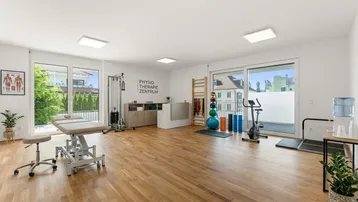 Heller Therapieraum mit Holzboden, großen Fenstern und Blick ins Grüne, ausgestattet für Physiotherapie.