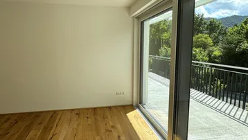Helles Zimmer mit Holzboden und großem Schiebefenster, das auf einen Balkon mit grüner Aussicht führt.