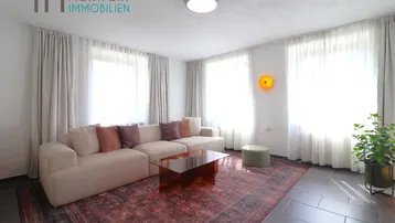 Helles Wohnzimmer mit beigem Sofa, modernem Teppich und großen Fenstern.