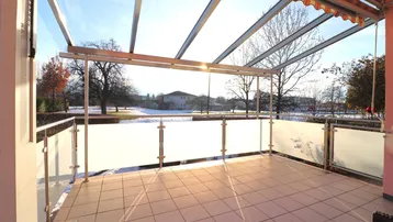 Überdachte Terrasse mit Fliesenboden und Glasgeländer, bietet einen weiten Blick ins Grüne.