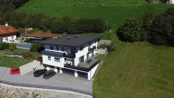 Modernes Gebäude mit Flachdach und Balkonen, eingebettet in eine grüne Hügellandschaft mit Bergblick.