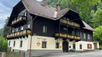 Traditionelles Gasthaus mit dunkler Holzfassade, mehreren Balkonen und Bergblick in grüner Umgebung.