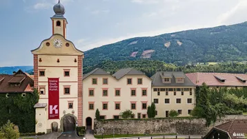 Historisches Gebäude mit markantem Turm und Bergkulisse, umgeben von üppiger Natur.