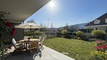 Sonnige Terrasse mit Gartenmöbeln, Rasenfläche und blühenden Pflanzen.