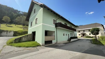 Grünes Wohnhaus mit Satteldach und Holzbalkonen in ländlicher Umgebung.