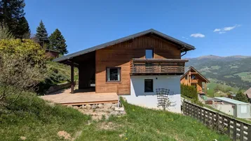 Charmantes Holzhaus mit großer Holzterrasse und Balkon, umgeben von grüner Natur und Bergblick.