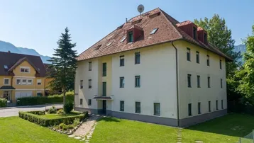 Mehrfamilienhaus mit Satteldach, Gaubenfenstern und gepflegter Grünfläche in bergiger Umgebung.