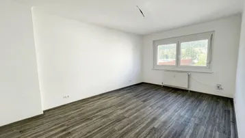 Helles, leeres Zimmer mit modernem Laminatboden und Fensterblick ins Grüne.