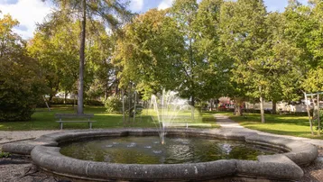Ein idyllischer Park mit einem Steinbrunnen und vielen Bäumen an einem sonnigen Tag.