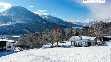 Winterlandschaft mit verschneiten Bergen und einem traditionellen Haus in alpiner Umgebung unter blauem Himmel.