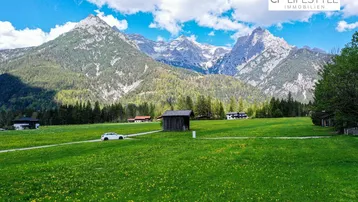 Weitläufige grüne Wiesenfläche mit traditioneller Holzhütte und majestätischem Bergblick unter blauem Himmel.