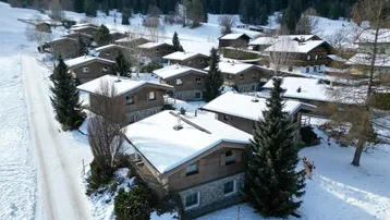 Luftaufnahme einer Gruppe von verschneiten Chalets in einer idyllischen Berglandschaft mit Wald im Hintergrund.