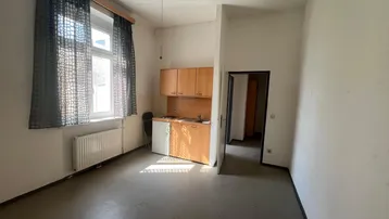 Heller Wohnraum mit kleiner Küchenzeile, großem Fenster und Deckenventilator.