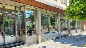 Ansicht der Geschäftsfassade mit großen Schaufenstern und Eingangsbereich, umgeben von Bäumen und Bürgersteig.