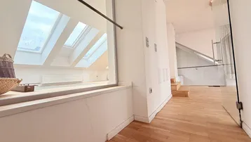 Heller Flur mit Holztreppe und Glasgeländer, der zu den oberen Etagen führt.