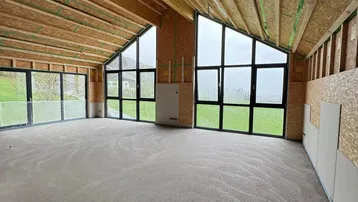Geräumiger, unfertiger Innenraum mit hohen Holzdecken und großen Fenstern mit Blick ins Grüne.