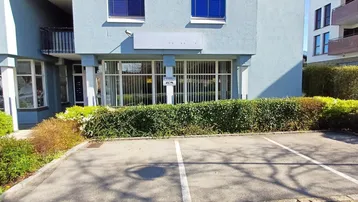 Gebäudefassade mit Geschäftsräumen im Erdgeschoss und einem Parkplatz davor, umgeben von einer Hecke.
