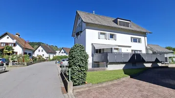 Weißes Wohnhaus mit Satteldach und Balkon in einer ruhigen Wohngegend mit viel Grün.