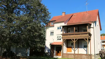 Altes Gebäude mit traditionellem Holzbalkon und großem Baum im Vorgarten.