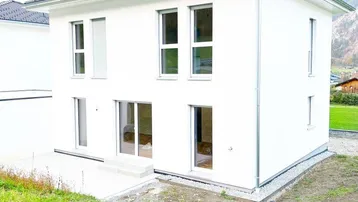 Modernes weißes Einfamilienhaus mit großen Fenstern und Bergblick in ländlicher Umgebung.