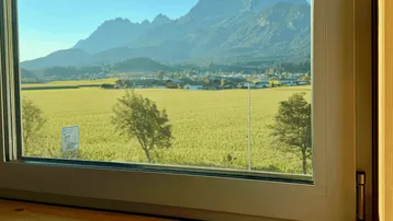 Panoramablick aus dem Fenster auf weite grüne Felder und beeindruckende Berge unter blauem Himmel.
