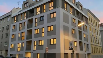 Modernes Mehrfamilienhaus mit beleuchteten Fenstern in der Dämmerung, umgeben von städtischer Architektur.
