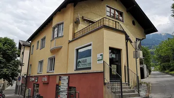 Dieses gepflegte Gebäude präsentiert sich mit einer traditionellen Architektur, einem Balkon und einem Geschäft im Erdgeschoss, umgeben von grüner Landschaft und Bergen.