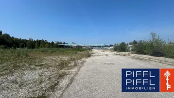 Weitläufiges unbebautes Grundstück mit Schotterweg und Blick auf entfernte Bäume unter blauem Himmel.