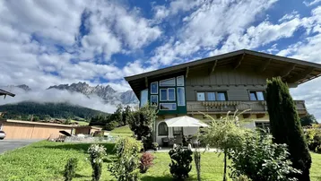 Charmantes Haus mit Terrasse und Garten, umgeben von einer malerischen Berglandschaft unter blauem Himmel.