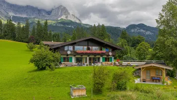 Großes Chalet-Haus in den Bergen mit grüner Wiese und beeindruckendem Alpenpanorama.