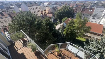Geräumiger Balkon mit Holzboden, Glasgeländer und weitem Blick über die Stadt und grüne Bäume.