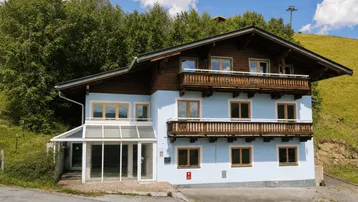 Traditionelles Haus im alpinen Stil mit hellblauer Fassade und Holzbalkonen, umgeben von grünen Hügeln.