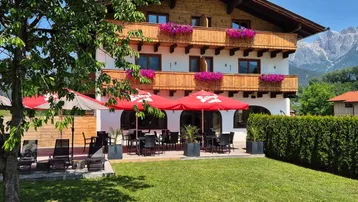 Traditionelles Gebäude mit Holzbalkonen, blühenden Blumenkästen und einer sonnigen Terrasse mit Garten.