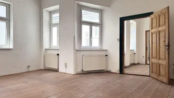 Geräumiger Raum mit Holzdielen, mehreren Fenstern und einer geöffneten Holztür zu einem Nebenraum.