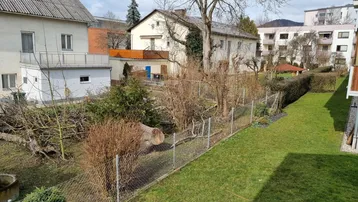 Grüner Gartenbereich mit Rasenfläche und Blick auf umliegende Wohnhäuser unter blauem Himmel.