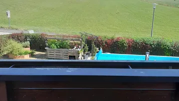 Weitblick vom Balkon über grüne Felder, Gärten und einen Pool in der Ferne.