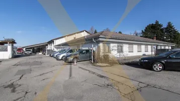 Gewerbegebäude mit Parkplätzen und asphaltierter Zufahrt unter blauem Himmel, das Renovierungsbedarf zeigt.