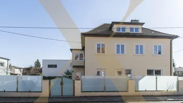 Vorderansicht des Hauses mit gepflegtem Vorgarten und moderner Zaunanlage unter blauem Himmel.