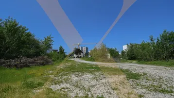 Blick über ein unbebautes Grundstück mit Schotterweg, umgeben von Bäumen, hin zu entfernten Gebäuden und einem Baukran unter blauem Himmel.