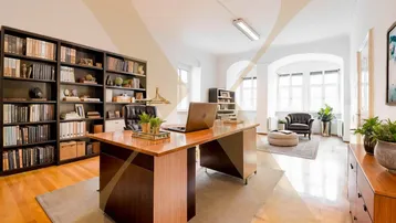 Geräumiges Büro mit Holzboden, großen Bücherregalen und viel Tageslicht durch die hohen Fenster.