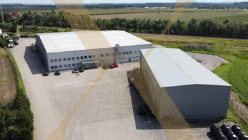 Luftaufnahme eines Industriegebiets mit zwei großen Hallen und umliegenden Feldern unter blauem Himmel.