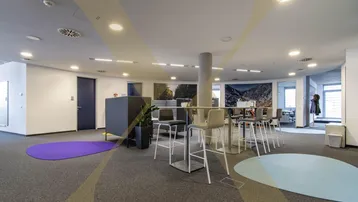 Ein moderner Gemeinschaftsbereich in einem Bürogebäude mit Stehtischen und farbigen Teppichinseln.
