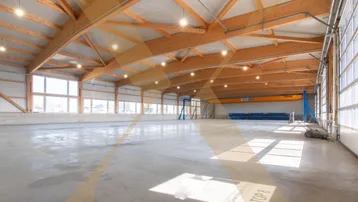 Geräumige Lagerhalle mit hoher Holzkonstruktion, Betonboden und viel Tageslicht durch große Fenster.