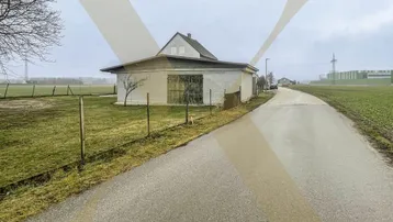Blick auf ein älteres Nebengebäude und das Hauptgebäude, umgeben von Feldern und einer Zufahrtsstraße.