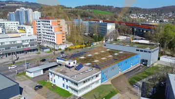 Luftaufnahme eines Gewerbegebiets mit mehreren Gebäuden, umgeben von Wohngebäuden und einer Brücke im Hintergrund.