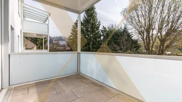 Heller Balkon mit Fliesenboden und Glasbrüstung, bietet einen Blick ins Grüne.
