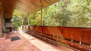 Geräumige überdachte Terrasse mit Fliesenboden, Holzbalken und integriertem Grillbereich, umgeben von viel Grün.