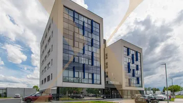 Modernes Bürogebäude mit Glasfassade und Parkplätzen unter einem bewölkten Himmel.