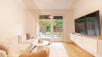 Helles Wohnzimmer mit beigem Sofa, Holzboden und Zugang zu einem kleinen Gartenbereich.