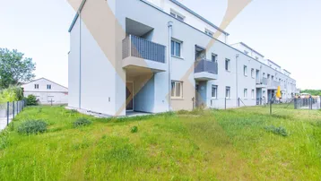 Modernes Mehrfamilienhaus mit Balkonen und einer Grünfläche im Vordergrund, umgeben von einer ländlichen Umgebung.